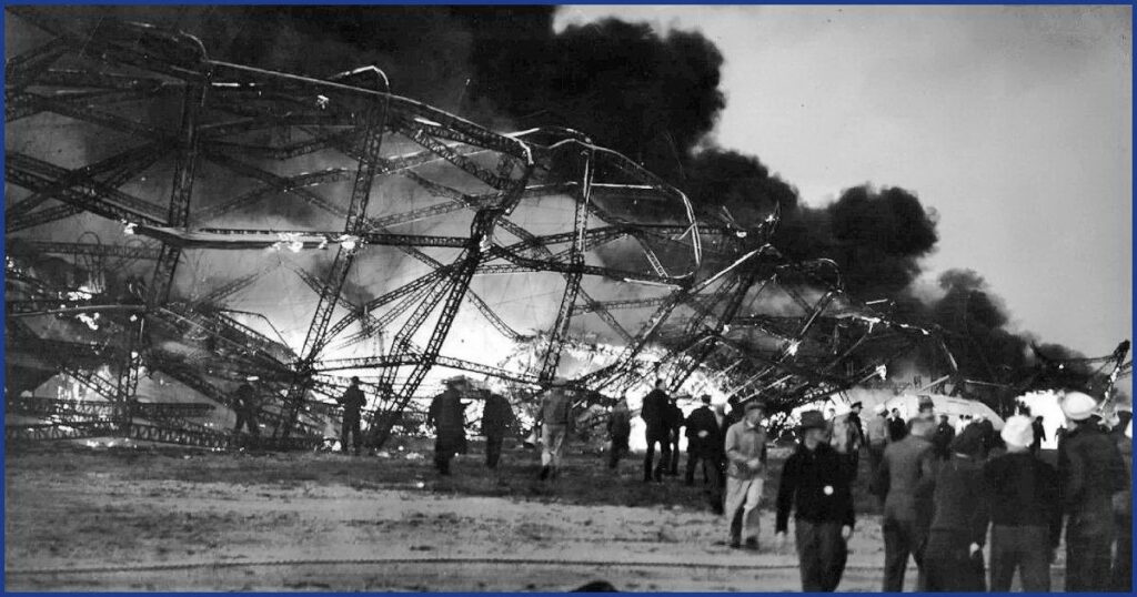 Чёрно-белая фотография: горящий каркас дирижабля Гинденбург на земле, вокруг дым и люди. На переднем плане лежит человек в форме.